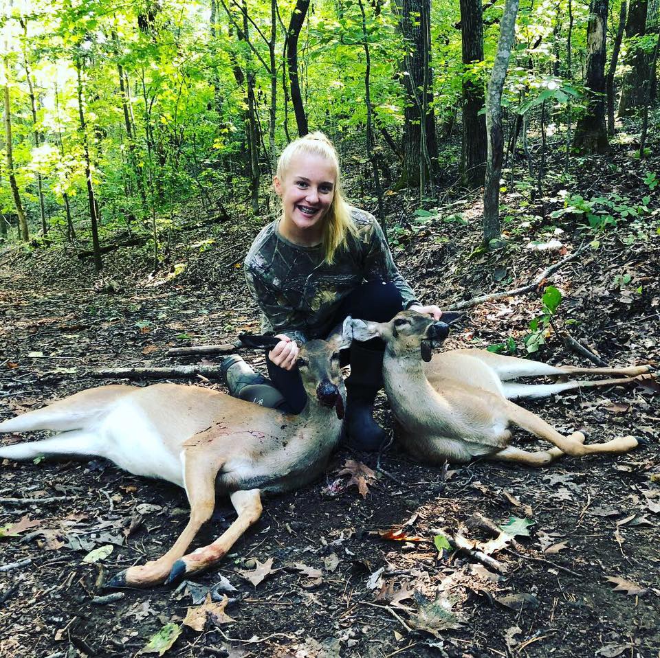 Young girl with blonde ponytail in arkansas woods with two whitetail doe that she harvested.